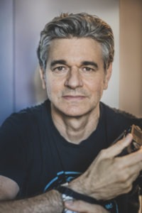 A middle-aged man with gray hair sits indoors, holding a camera. He is wearing a dark t-shirt and has a neutral expression on his face. The background is softly blurred, focusing attention on the man, who appears to be immersed in his Berkeley Journalism work.