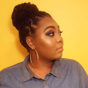 A person with braided hair styled in a bun is looking to the side, wearing a denim shirt and large hoop earrings. The individual, who appears to be a Berkeley Journalism student, has dramatic eye makeup and a subtle, glossy lip. The background is a solid yellow color.