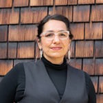 A person with glasses, long earrings, and a nose piercing is standing against a background of wooden shingles. They are wearing a black top and a grey vest, with their hair tied back—perfectly capturing the eclectic vibe of Berkeley Journalism.