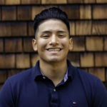 A person with short, dark hair styled upwards and wearing a navy blue polo shirt is smiling in front of a wooden shingle wall, exuding the confidence often seen in Berkeley Journalism graduates.