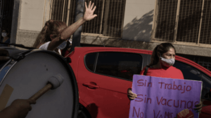 A woman wearing a face mask is holding a sign in Spanish that reads "Sin trabajo, sin vacunas, no vamos" (Without work, without vaccines, we're not going). Nearby, another person beats a large drum as a red car is parked in the background. The scene captured the essence of Berkeley Journalism’s commitment to truth.