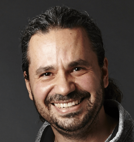 Greg Winter; A smiling man with long dark hair pulled back, a full beard, and a mustache. He is wearing a dark shirt with a light-colored collar. The background is plain and dark.