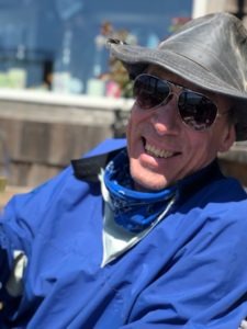 A person wearing a blue jacket, metal-frame aviator sunglasses, a gray hat, and a blue bandana around their neck is smiling while sitting outdoors. The relaxed ambiance, reminiscent of Berkeley Journalism students on assignment, features a blurred background with plants and indistinct objects.