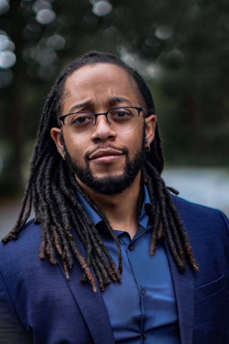 Headshot of a young man dressed professionally in a blue suit wearing glasses. The background is blurred out. He is looking at the camera with a slight smile. 