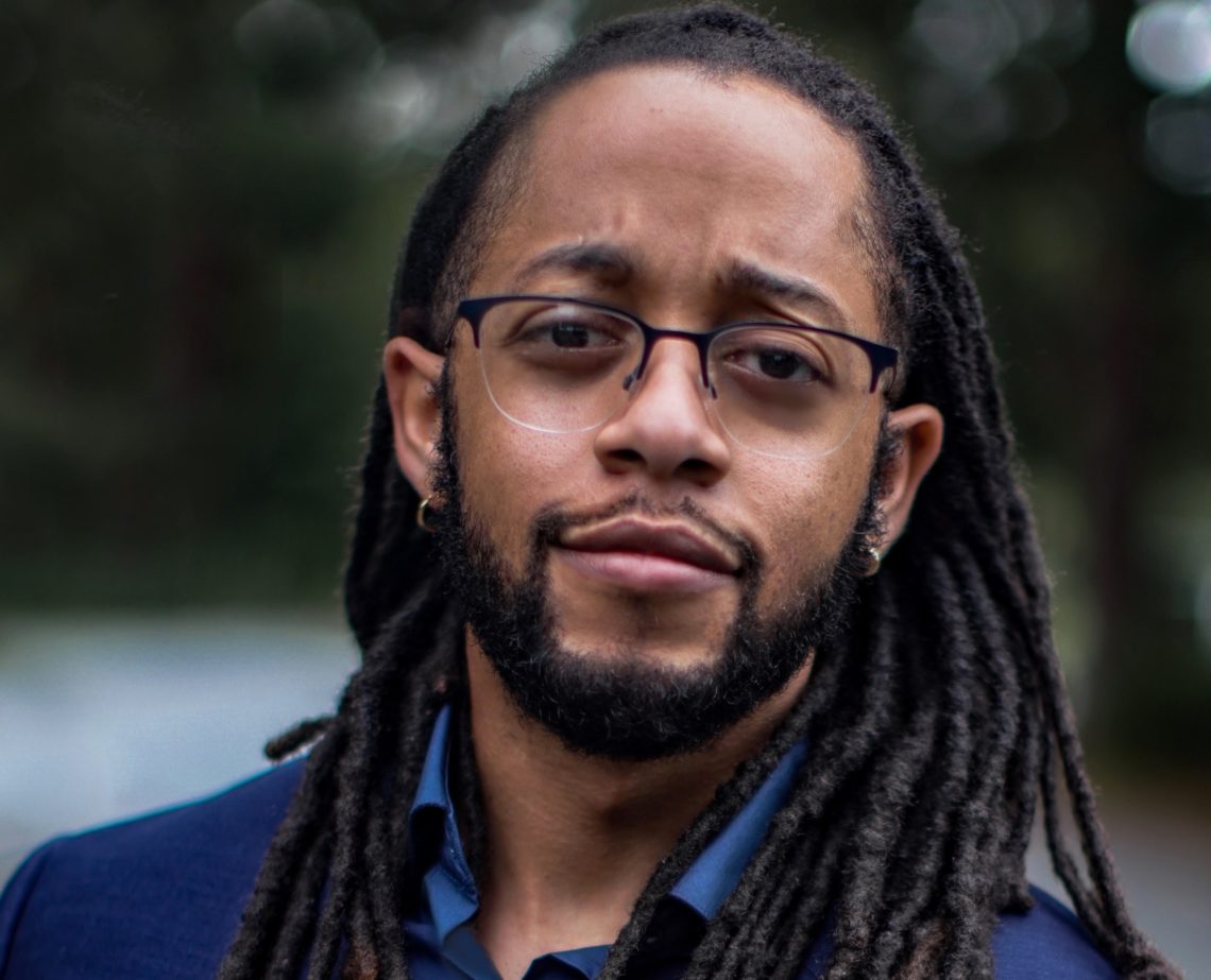 A person with long, dark dreadlocks, a beard, and wearing glasses, is looking at the camera with a thoughtful expression. Dressed in a dark blue blazer, they convey the intellectual vibe often seen in Berkeley Journalism circles. The background is blurred greenery, suggesting an outdoor setting.