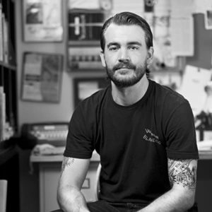 A bearded man with tattoos, wearing a dark T-shirt, sits in a cluttered office space filled with papers and documents pinned on the wall behind him. He has a calm expression and is looking at the camera. The black-and-white image evokes a Berkeley Journalism vibe, capturing raw storytelling essence.
