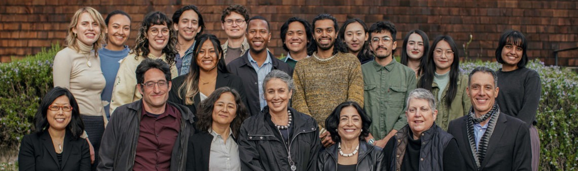 A group of 22 people stands close together, smiling and posing for a photo outdoors. They range in age and ethnicity, dressed in casual to business-casual attire. The camaraderie reflects the diverse spirit of Berkeley Journalism, with a backdrop of a brick wall and greenery adding charm to the scene.