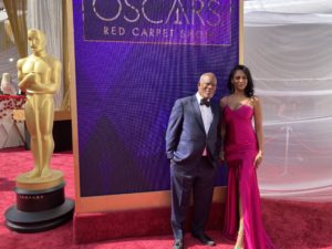 Two individuals, a man in a black tuxedo and a woman in a magenta gown, stand on the red carpet at the Oscars. They pose beside a large backdrop with "Oscars Red Carpet Show" displayed and a gold Oscar statue. The scene, perfect for Berkeley Journalism students to capture, is set against draped white fabric.