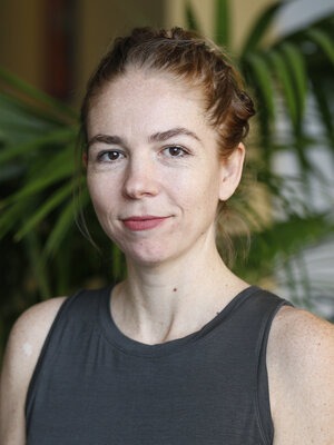 A woman with light skin and auburn hair pulled back in a braided updo stands poised, her sleeveless gray top contrasting beautifully with the blurred green background of broad-leafed plants, exuding the calm confidence often seen in Berkeley Journalism profiles.