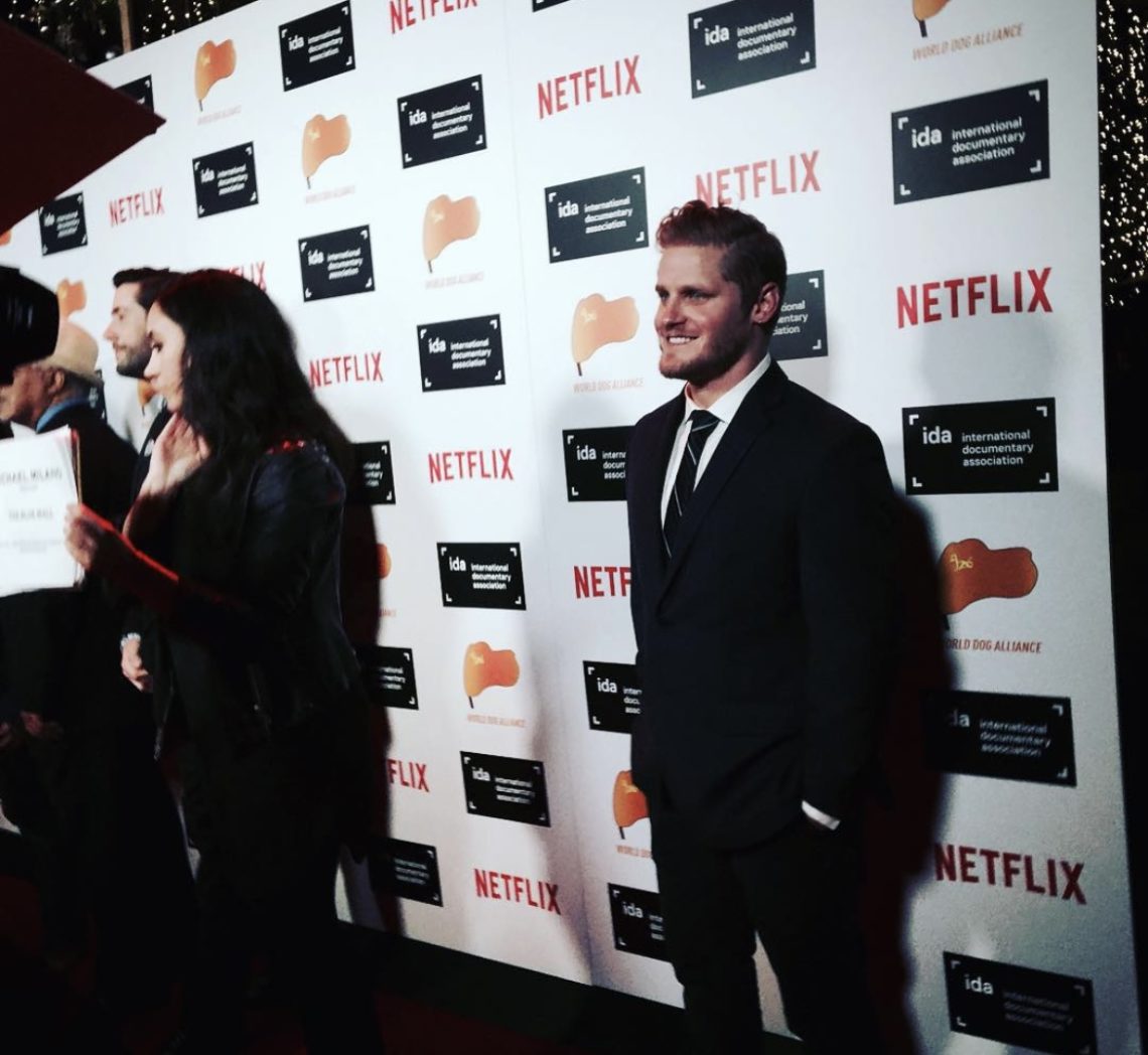 A man in a suit stands on a red carpet with a step-and-repeat backdrop displaying logos for Netflix, Berkeley Journalism, and the IDA (International Documentary Association). A woman holds a sign in the foreground, partially obscuring other individuals. The setting includes bright event lighting.