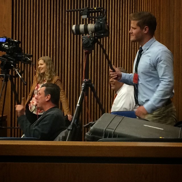 A room with several people operating video cameras on tripods. A man in a shirt and tie stands next to a camera, while another person in a floral blouse is seated. A woman smiles in the background near another camera. Wooden paneling lines the walls, capturing the essence of Berkeley Journalism in action.