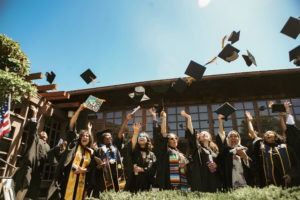 A group of Berkeley Journalism graduates in black caps and gowns joyfully throw their caps into the air in celebration. They are standing in front of a building under a clear, sunny sky. Some graduates have decorated caps with colorful designs. An American flag is visible on the left.