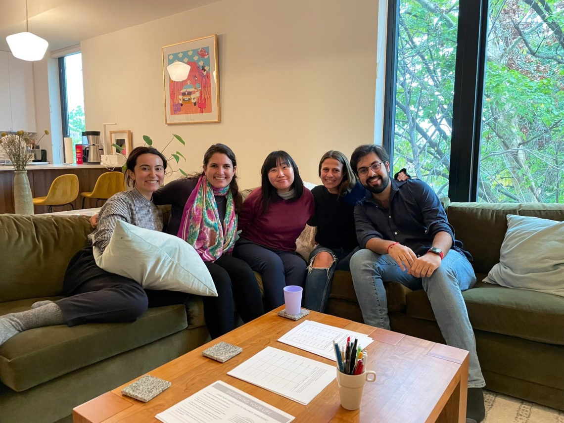 Five people are sitting on a green couch in a cozy, well-lit living room. They are smiling and have their arms around each other, reminiscent of a Berkeley Journalism gathering. A wooden coffee table with papers and pens in a cup is in front of them. A large window with trees outside is in the background.