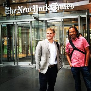Two men stand in front of The New York Times building entrance. The man on the left wears a light grey blazer and white shirt, while the man on the right, perhaps a proud Berkeley Journalism alumnus, dons a red short-sleeve button-down shirt and carries a shoulder bag. Both are smiling and relaxed.