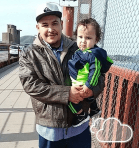 A man named Adam Solorzano, wearing a brown jacket, blue jeans, and a grey cap, is standing on a bridge, holding a baby dressed in a green and blue tracksuit. They are next to a chain-link fence with a cloudy sky icon in the corner of the image.