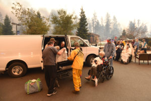 Search and rescue team and a person in scrubs help transfer three older women in to a van, in the background smoke from the Camp fire can be seen as the Feather River hospital burns behind them.