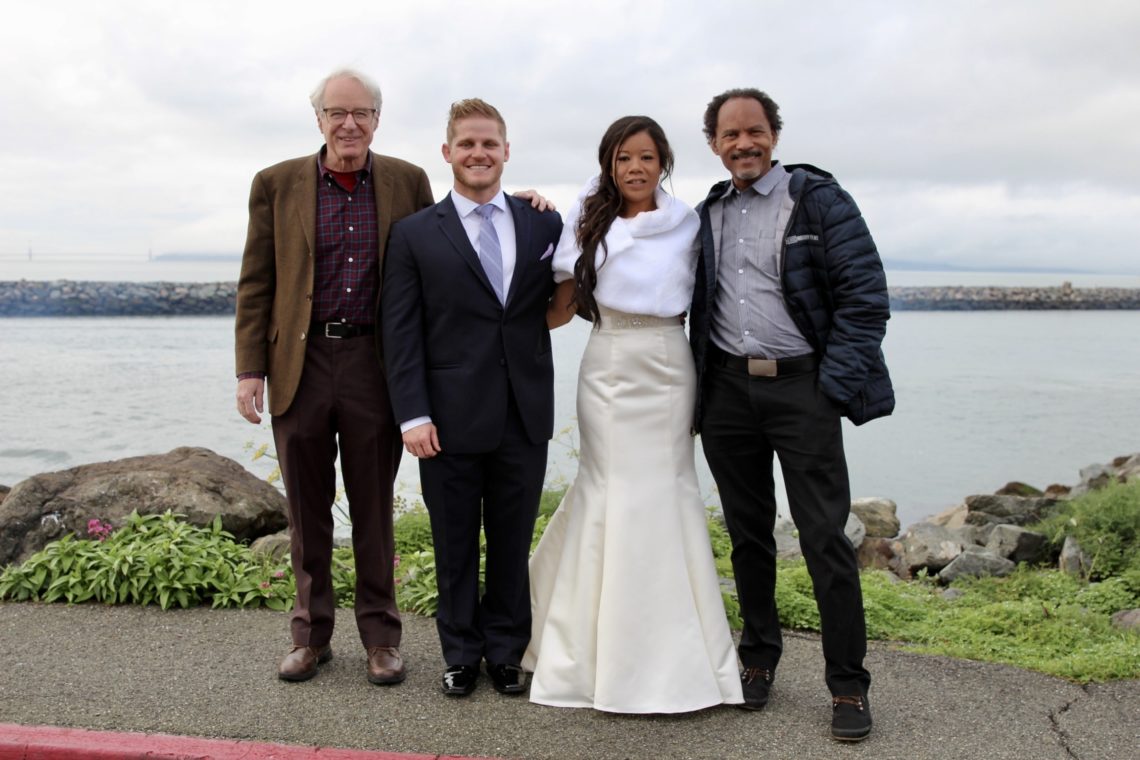 Four people stand together by a rocky shoreline, smiling for a photo. One woman is wearing a white formal dress while the three men are dressed in formal and semi-formal attire. The ocean and a cloudy sky are visible in the background, capturing a picturesque moment worthy of Berkeley Journalism.