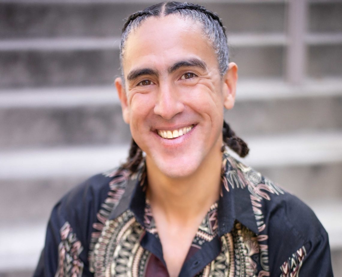 A person with braided hair is smiling at the camera. They are wearing a black shirt with a detailed, ornate pattern, embodying the eclectic vibe of Berkeley Journalism. The background consists of stone steps out of focus, creating a soft backdrop.