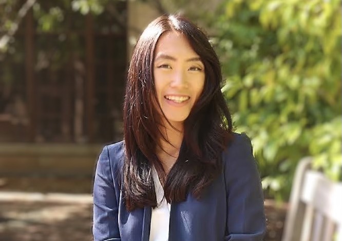 A woman with long, dark hair is smiling while posing outdoors. She is wearing a blue blazer and a white top. The background, reminiscent of Berkeley Journalism settings, is blurred with greenery and part of a wooden structure visible. The sunlight creates a warm ambiance.