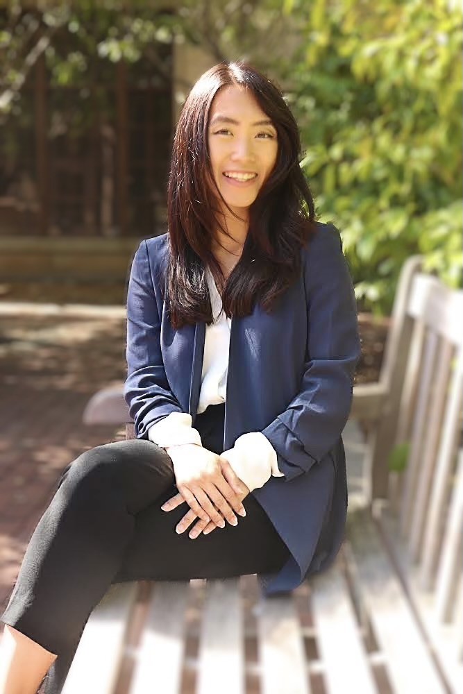 A woman with long dark hair is sitting on a wooden bench outdoors. She is smiling at the camera, wearing a navy blazer over a white blouse and black pants. The background includes greenery and what appears to be a building structure, hinting at the serene environment of Berkeley Journalism.