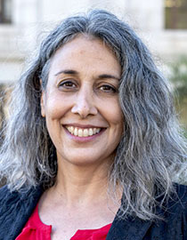 A person with long, gray hair and a bright smile is pictured outdoors, their black jacket over a red top. The background, slightly blurred with a building and some greenery, evokes the serene atmosphere around Berkeley Journalism.