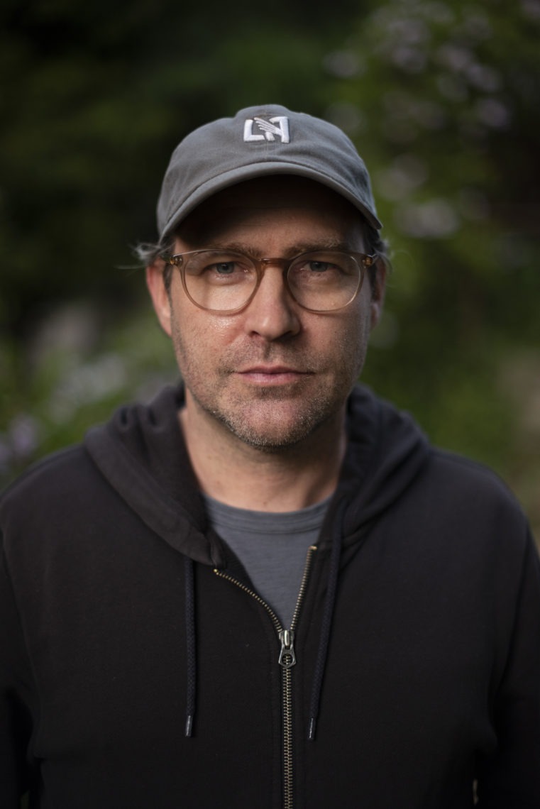 Jeffrey Plunkett. A middle-aged person with slight stubble is seen wearing glasses, a cap, and a dark hoodie. The background is blurred out, showcasing greenery. The individual has a neutral expression and is looking directly at the camera.