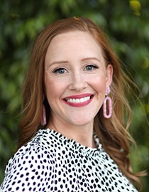 A woman with long, wavy auburn hair is smiling at the camera. She is wearing pink oval-shaped earrings and a white dress with black polka dots. Against a backdrop of blurred greenery, she exudes the effortless grace and confidence often seen in Berkeley Journalism students.