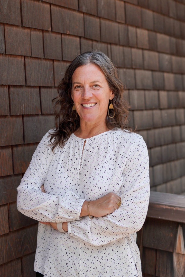 A middle aged woman wearing a long white blouse, smiling, and standing with her arms crossed in front of a shingled wall.