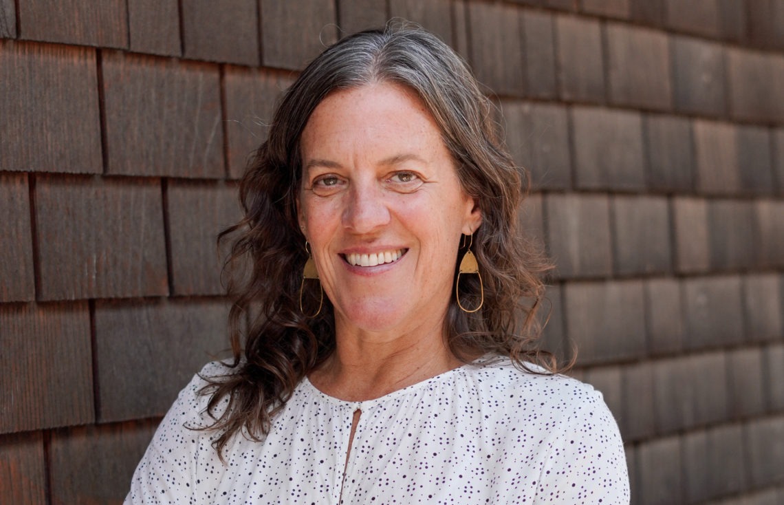 A woman with long, wavy hair and wearing a white blouse smiles at the camera. She is standing in front of a wooden shingle wall, wearing gold geometric-shaped earrings. This new hire is excited to join the team led by the person who runs Berkeley Journalism.