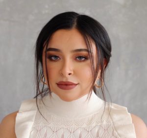 A woman with dark hair parted down the middle and loose strands framing her face is wearing gold hoop earrings and a white sleeveless turtleneck top with lace details. She stands against a plain, textured gray background, her serene expression reminiscent of farmworkers in peaceful moments.