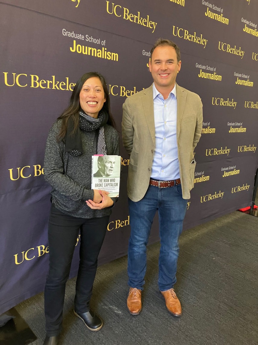 Two people, a man and a woman, standing next to each other smiling at an event. There is a Graduate school of Journalism backdrop. The woman is holding a book titled, "The Man Who Broke Capitalism". 