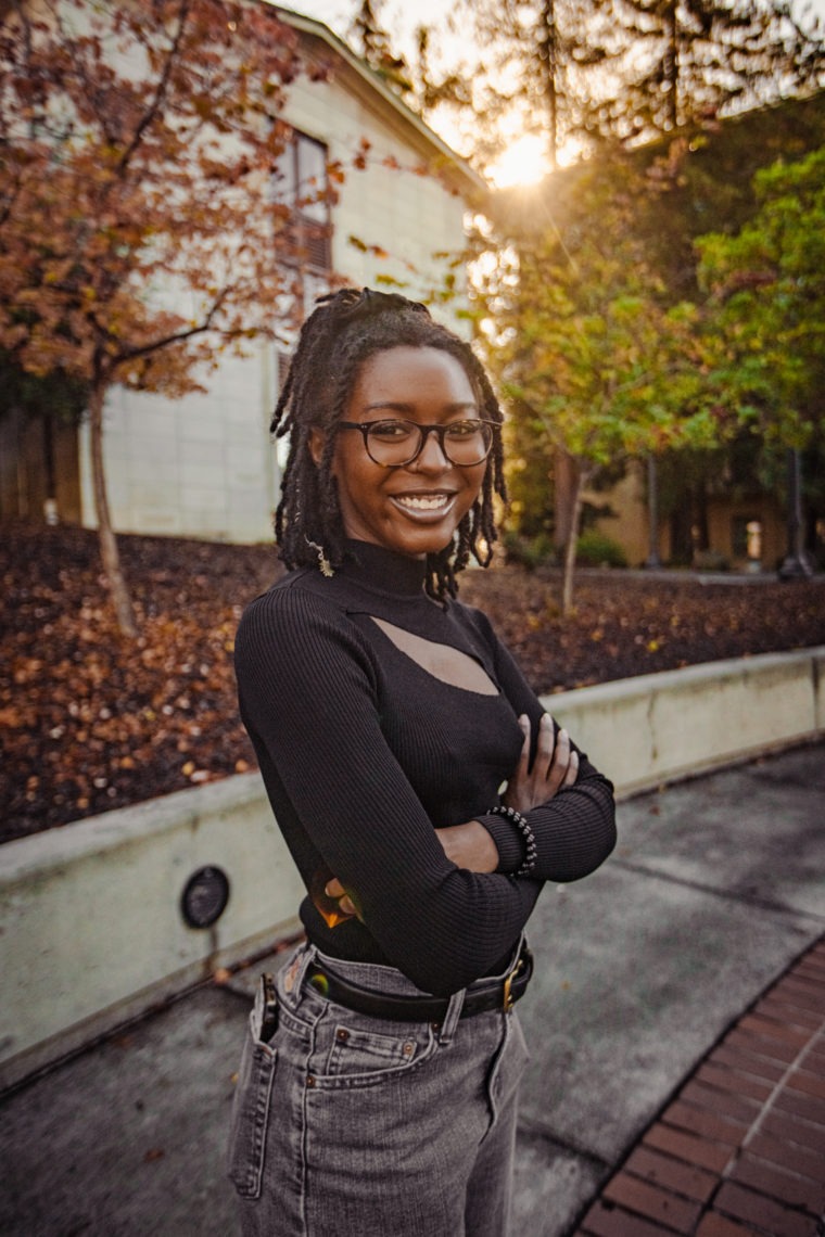 Photo of an African American woman with a cool black top and grey jeans on wearing braids and glasses with her arms crossed smiling.