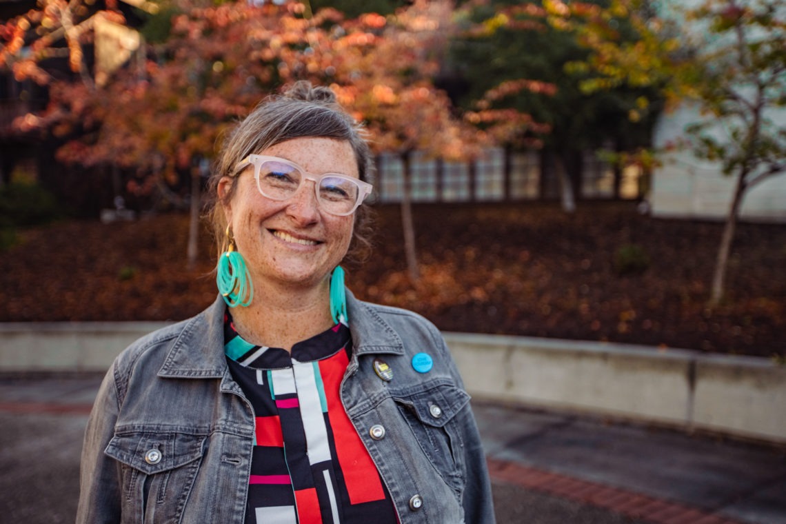 Photo of a women with her light brown hair up in a barret wearing pink glasses, big turquoise round earrings and red white and black patterned top and a grey denim jacket smiling.