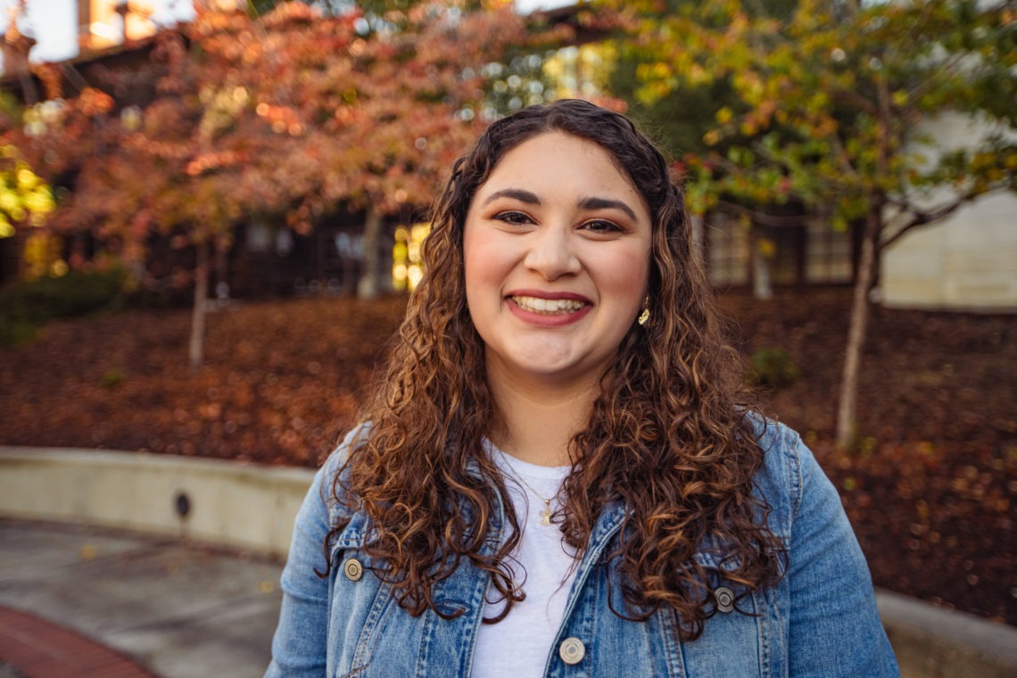 Photo of a Hispanic woman with long brown curly hair wearing a jean jacket over a white shirt smiling in front of the school on a fall day.