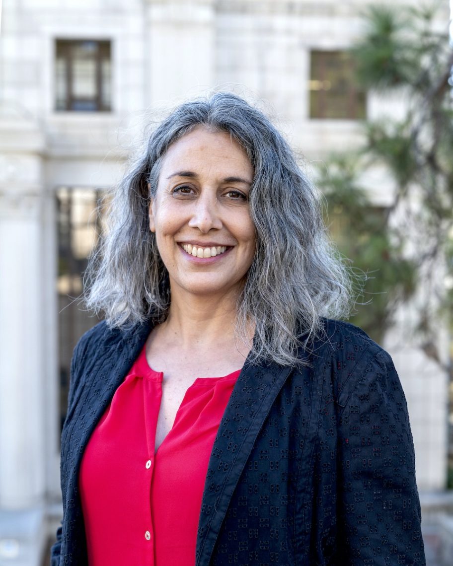 A woman with long, wavy gray hair smiles at the camera. Geeta Anand is wearing a black textured blazer over a bright red blouse. The background shows a building with classical architectural elements and some greenery. She recently received the Richard J. Levine Journalism Champion Award from the Dow Jones News Fund.