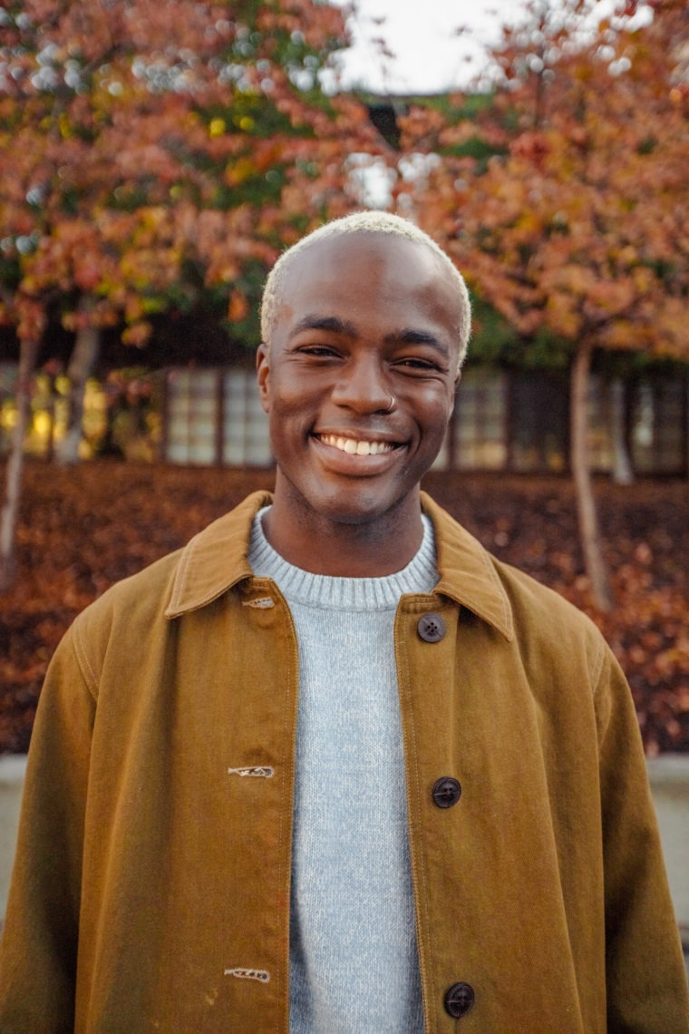 Photo of an African American man with bleached short hair wearing an elegant camel colored overcoat over a grey crew neck sweater in front of fall foliage smiling.