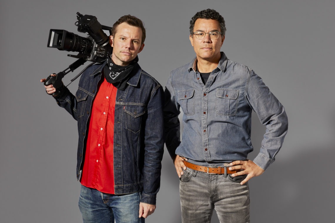 Two people standing against a gray background. The person on the left is holding a camera on their shoulder and wearing a denim jacket over a red shirt. The person on the right, featured in our Quarterly Newsletter, is wearing glasses, a blue shirt, and placing one hand on their hip.