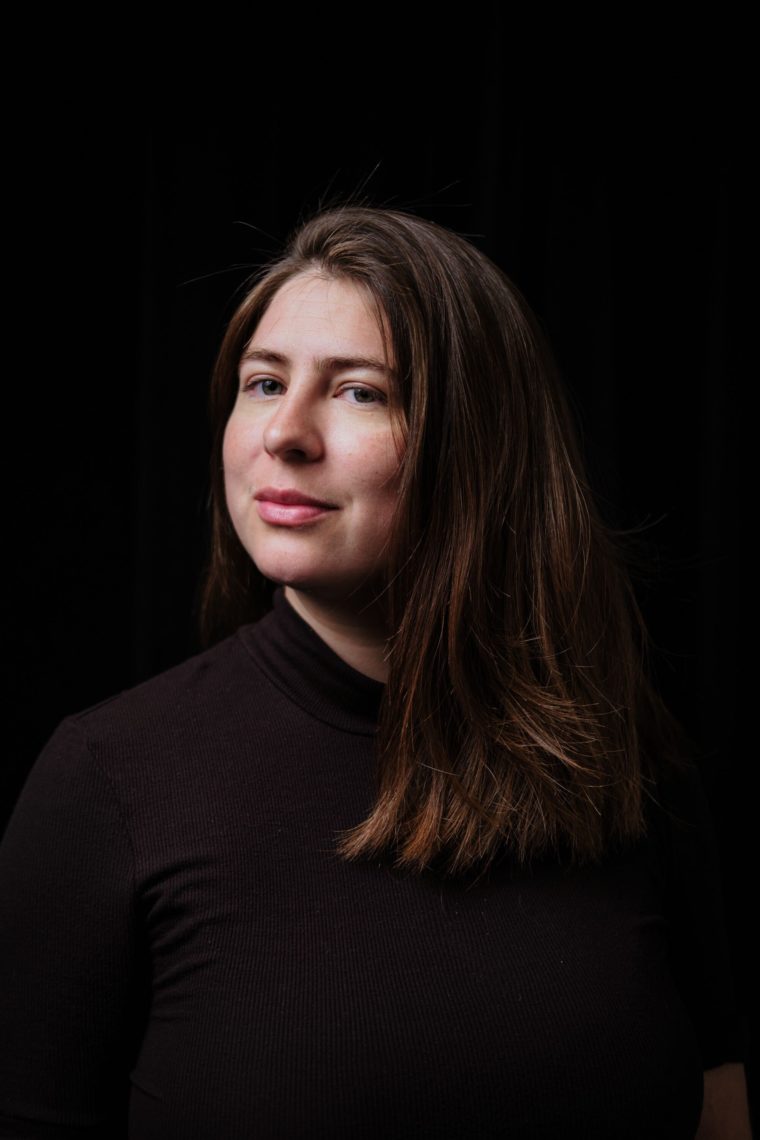 A woman with shoulder length hair is photographed. She is looking sideways and the black sweater she is a wearing blends into the black background of the photo. 