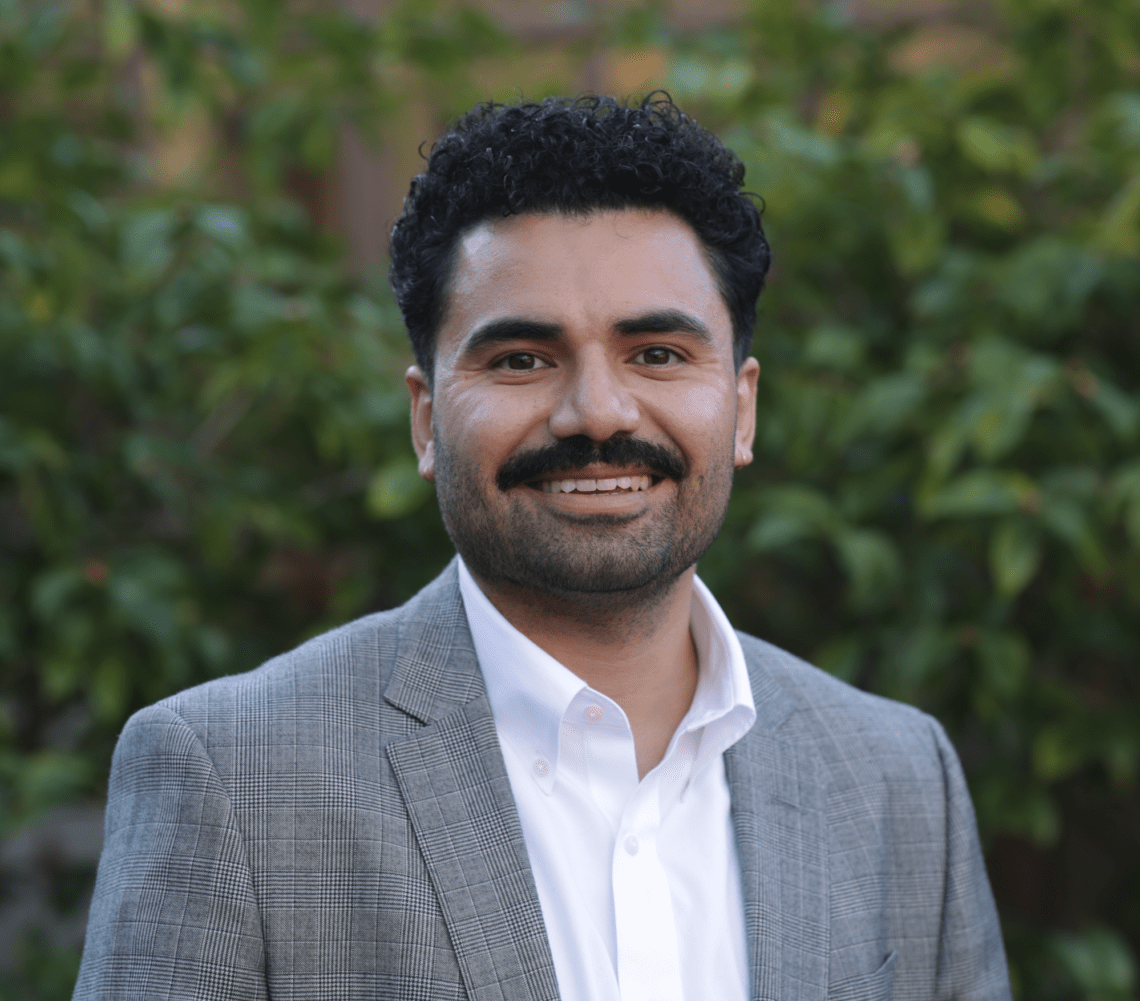 Daniel Márquez, an assistant director, is smiling at the camera with his curly hair and mustache. He is wearing a gray plaid blazer over a white shirt, standing against a backdrop of green foliage.