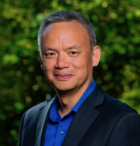 Edward Wong, a man with short gray hair, is dressed in a dark suit and blue shirt. He is smiling against a blurred green background.