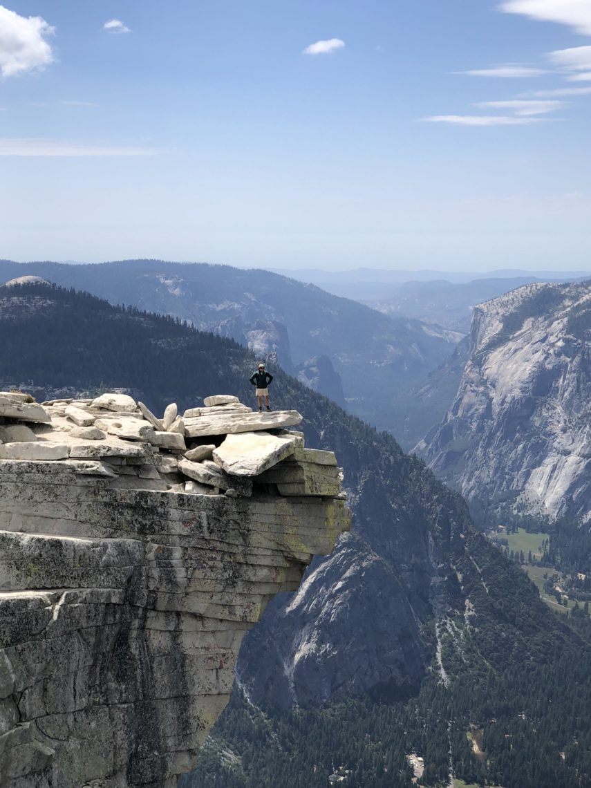 A lone hiker stands on the edge of a rocky cliff, overlooking a vast mountain landscape with valleys and peaks stretching into the distance under a partly cloudy sky. The scene, reminiscent of mesmerizing documentary filmmaking, emphasizes the grandeur of nature.