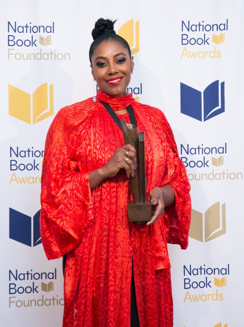 A woman is standing in front of an event banner that says, "National Book Foundation". Her hairstyle is in a bun, she is wearing earrings, and has on a long lavish red dress. 