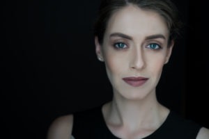 A woman with fair skin and light blue eyes gazes into the camera, against a black background. She has neatly styled brown hair and is wearing subtle makeup, including nude lipstick. Representing women filmmakers, she wears a sleeveless black top and small stud earrings.