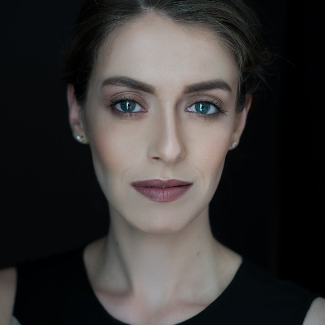 A woman with light skin and blue eyes, embodying the strength of women filmmakers, is looking directly at the camera. She is wearing a dark sleeveless top, subtle makeup, and small stud earrings. The black background makes her features stand out prominently.