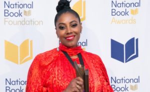 A woman wearing a vibrant red outfit smiles while holding an award. She stands in front of a backdrop with the "National Book Foundation" and "National Book Awards" logos, celebrating her win for The Yellow House, a notable work of nonfiction.