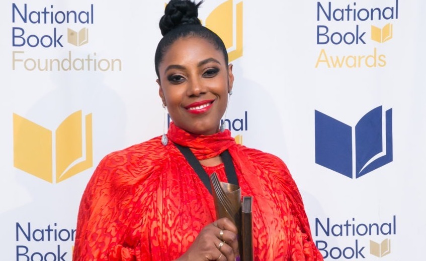 A woman wearing a vibrant red outfit smiles while holding an award. She stands in front of a backdrop with the "National Book Foundation" and "National Book Awards" logos, celebrating her win for The Yellow House, a notable work of nonfiction.