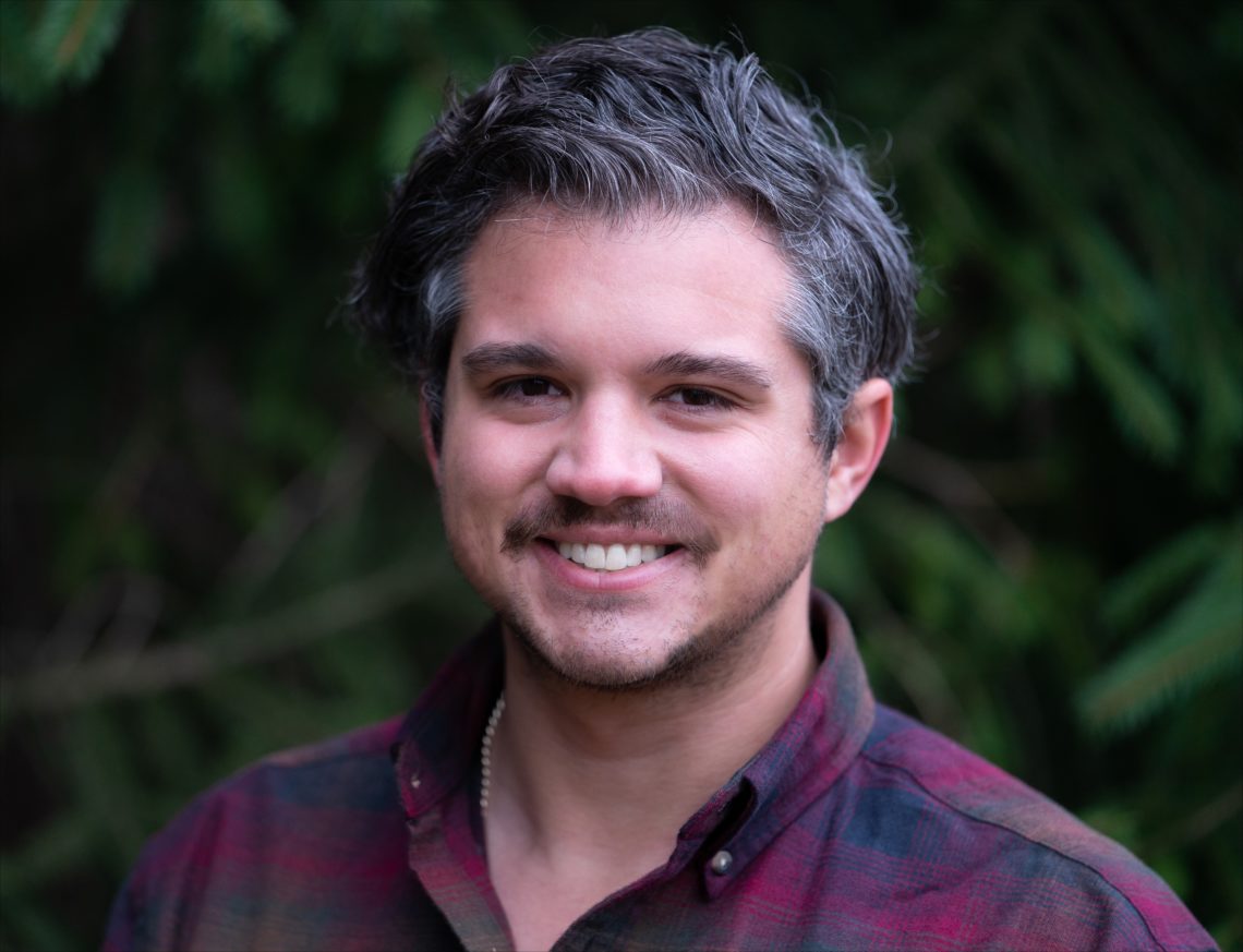 A man with short, wavy, salt-and-pepper hair and a beard is smiling at the camera. He is wearing a maroon plaid shirt and standing in front of a blurred natural background with green foliage. This image of Chris Ehrmann captures his casual charm as he delves into the world of documentary filmmaking.