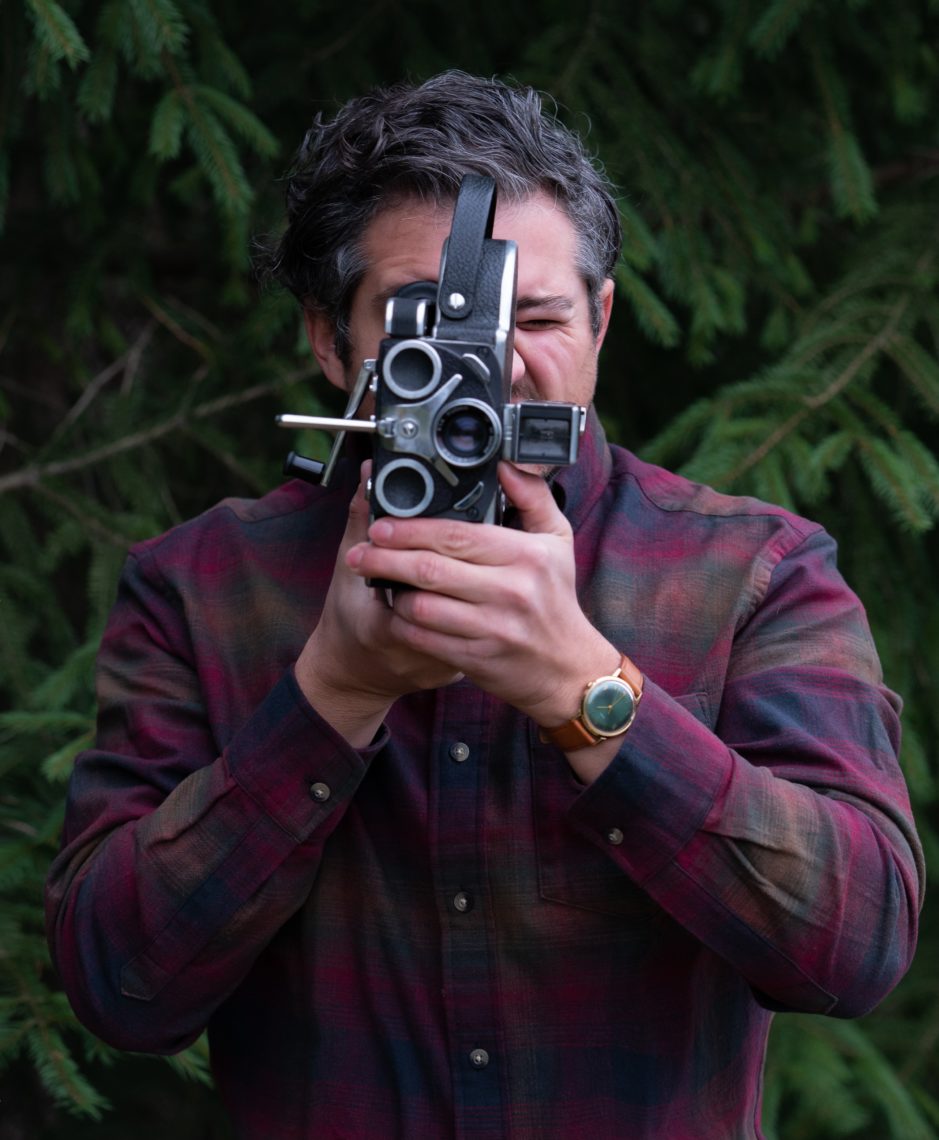 A man is looking into a film camera. He is wearing a red plaid shirt, a watch on his left wrist and is standing outside. The background is leaves and branches. 