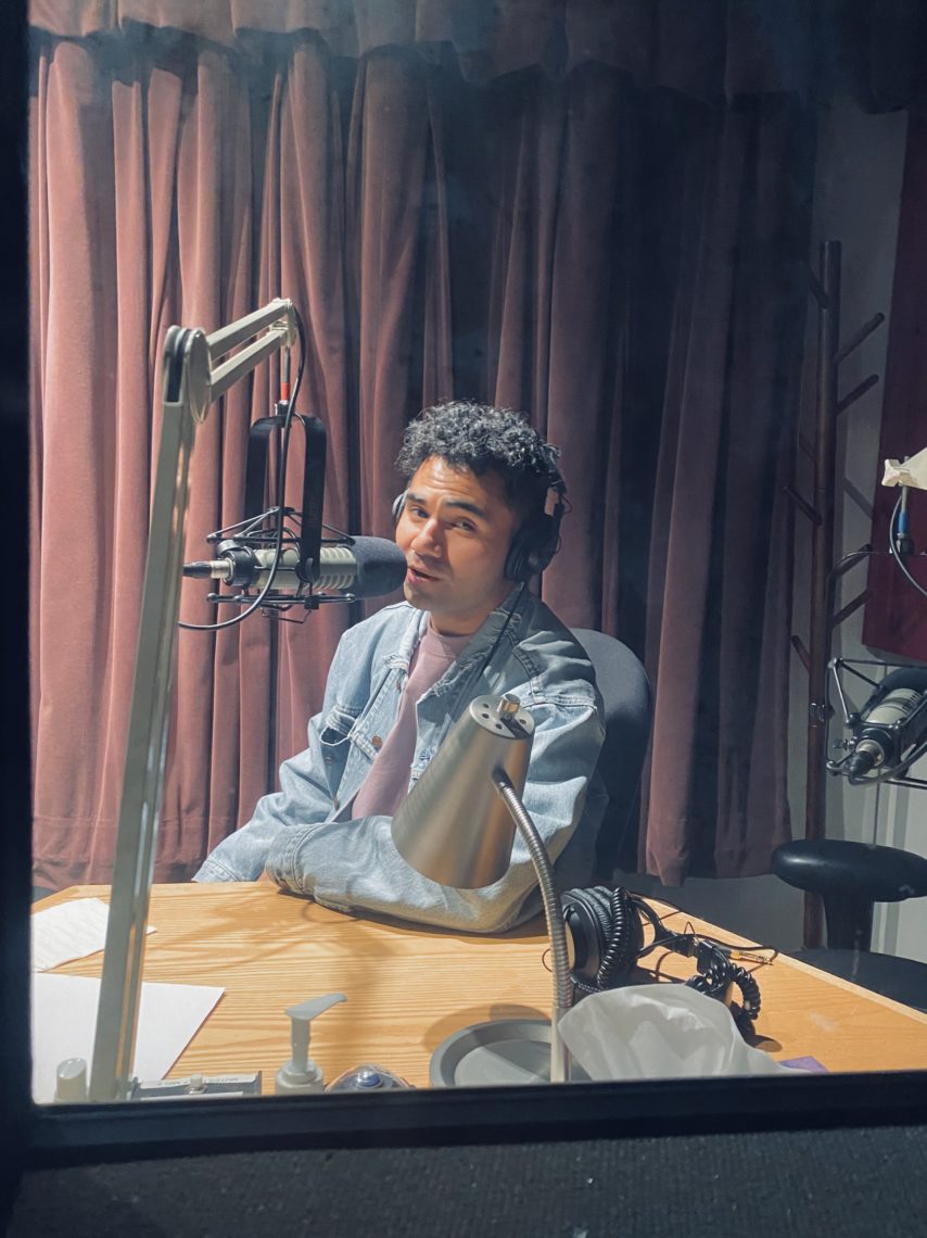 A young man has on headphones and is speaking into a microphone, recording an episode in a studio.