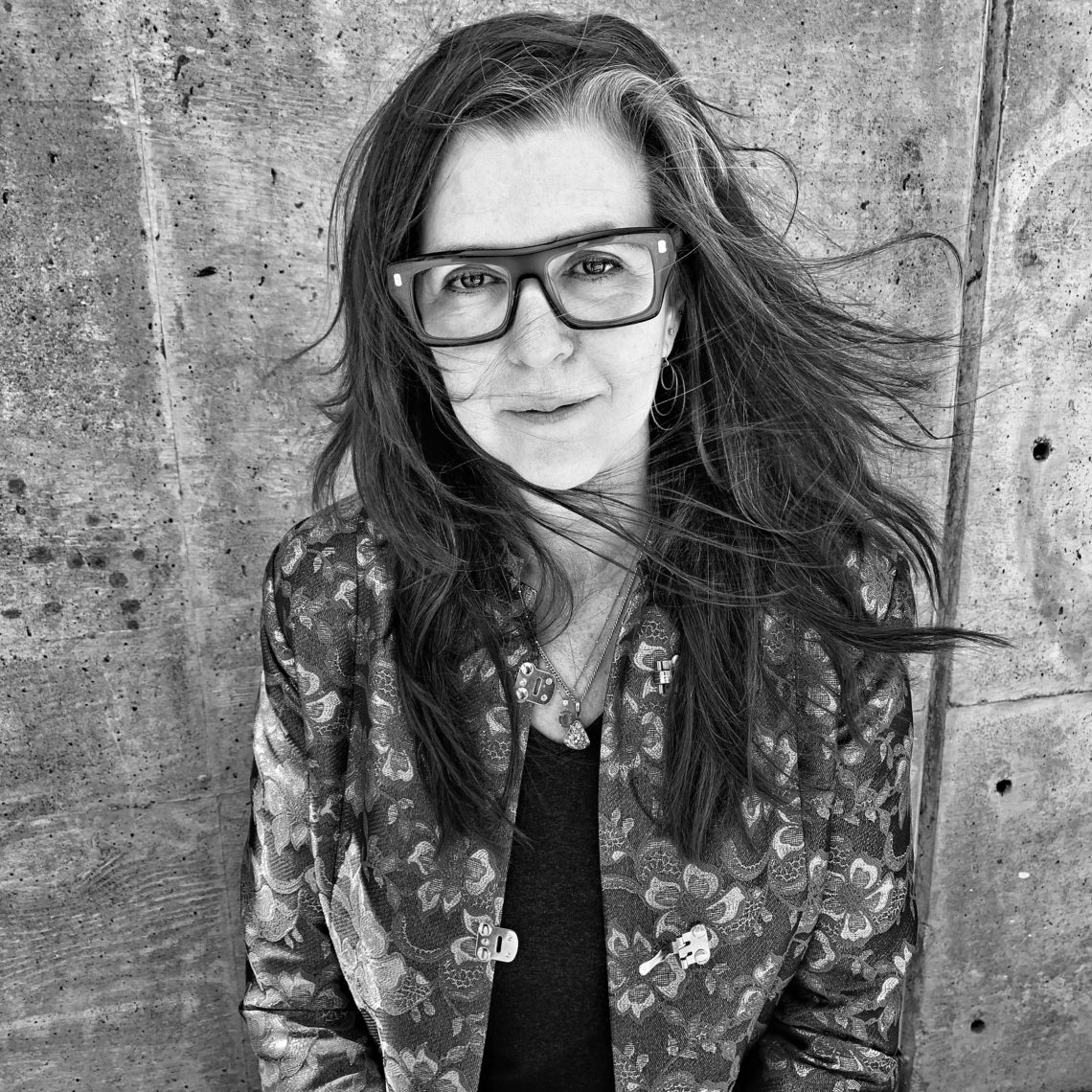 Black and white photograph of photographer Nigel Poor standing in front of a cement background with long brown hair blowing in the wind, wearing black glasses and a printed jacket.